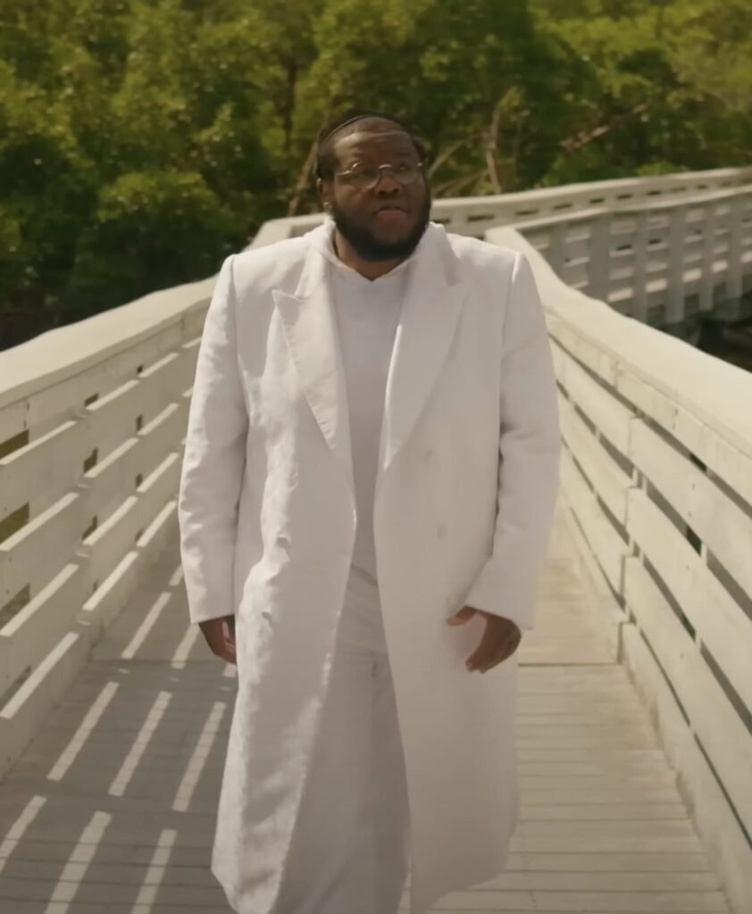 Man in a white suit and long overcoat walking along a white wooden boardwalk through trees